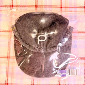 May include: A dark gray denim baseball cap with a white embroidered "p" logo, sealed in a clear plastic bag. The cap is folded and appears new. The background is a pink and red plaid pattern.
