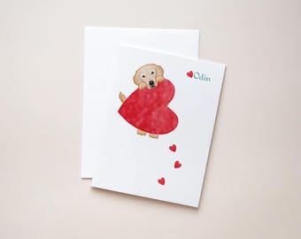 May include: A white card with a golden retriever puppy holding a red heart. The puppy is looking at the heart. The card has the name "Odin" written on it in blue text. There are five small red hearts scattered around the large heart.