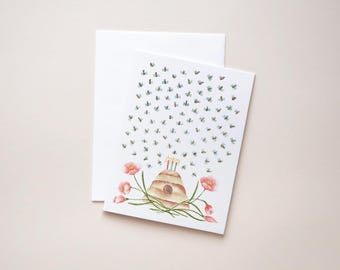 May include: A greeting card with a watercolor illustration of a beehive with two lit candles, surrounded by bees and pink flowers. A white envelope is behind the card. The card is on a light pink surface.