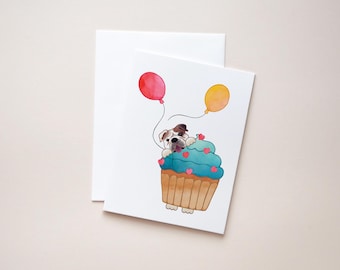 May include: A white bulldog wearing a blue and white cupcake hat with pink hearts. The bulldog is holding two balloons, one red and one yellow.