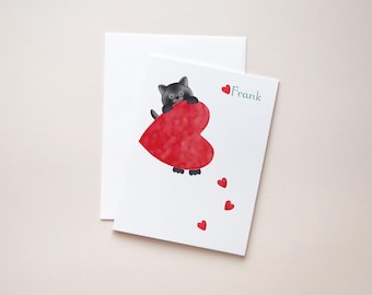May include: A white greeting card featuring a cartoon illustration of a grey cat holding a large red heart. The name Frank is printed in teal next to a small red heart, with three additional small red hearts floating below the cat.