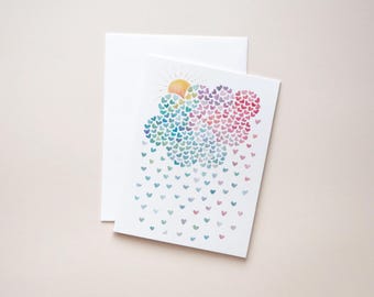 May include: A white greeting card and matching envelope featuring a watercolor illustration of a cloud made of small, multicolored hearts in shades of blue, purple, and pink, with a yellow sun peeking out and heart-shaped raindrops falling below.