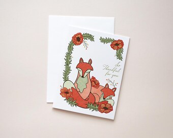 May include: A greeting card featuring two foxes with red-orange fur, nestled together. The card is decorated with red flowers and green foliage, with the words "Be Thankful for you" in script. A white envelope is behind the card.