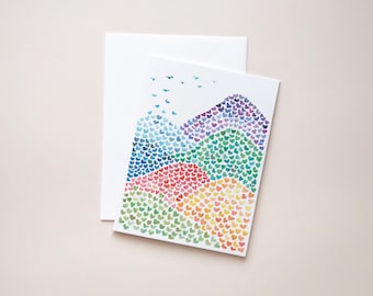 May include: A greeting card with a watercolor painting of colorful mountains made of hearts. The mountains transition from blue to green, red, and yellow. A white envelope is behind the card, and small blue birds fly above the mountains.