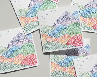 May include: A collection of square greeting cards featuring a mountain landscape design. The mountains are composed of small, colorful hearts in shades of red, orange, yellow, green, blue, and purple. Small bird silhouettes are scattered across the top of the cards.