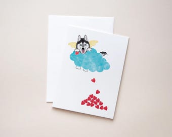 May include: A white greeting card with a watercolor illustration of a Husky dog with angel wings on a blue cloud, holding a red heart. Red hearts fall from the cloud. A white envelope is behind the card.