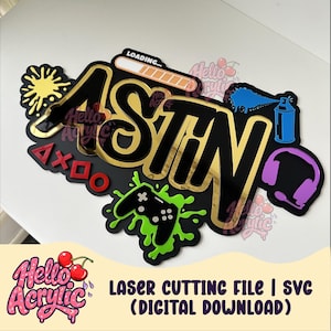 May include: A custom name sign featuring the name ASTIN in black and gold lettering, topped with a loading bar. The design includes a yellow paint splatter, red gaming controller symbols, a green controller, a blue spray can, and purple headphones.