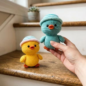 May include: Two crocheted ducklings are displayed on a wooden surface. One is yellow with an orange beak and feet, and a pink and white striped hat. The other is teal with an orange beak and feet, and a blue and white striped hat.