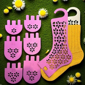 Peut inclure: Ensemble de gabarits roses pour chaussettes, avec des découpes florales, tailles 26-27 à 32, accompagnés d'une chaussette jaune et d'une forme de chaussette rose. Sur un fond vert parsemé de marguerites.