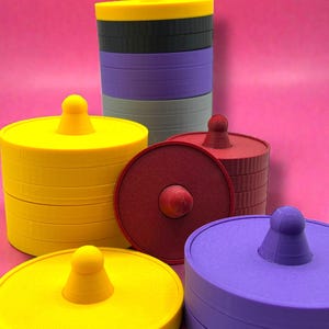 Könnte beinhalten: Eine Sammlung farbenfroher, runder Objekte mit einem zentralen Knopf. Die Objekte sind in Gelb-, Rot-, Lila-, Grau- und Schwarztönen gehalten. Der Hintergrund ist einfarbig rosa. Die Objekte scheinen Deckel oder Behälter zu sein.