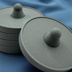 May include: A stack of gray coasters with a textured surface and a central knob. One coaster is separated from the stack, showing the design. The coasters are made of a solid material and are designed to protect surfaces.