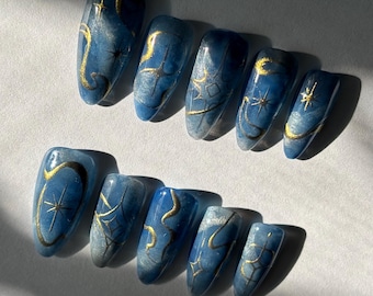 May include: A set of ten blue and gold nail designs. Each nail features a different celestial design, including stars, moons, and abstract gold lines. The nails have a glossy finish and are arranged on a white surface.