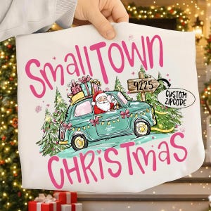 May include: White Christmas-themed tote bag with the words "Small Town Christmas" in pink. Santa Claus drives a teal car carrying gifts. The image includes a custom zip code and Christmas tree accents.