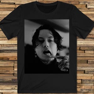 May include: Black t-shirt featuring a black and white portrait. The portrait shows a person with dark hair, tattoos, and an open mouth, with a finger in their mouth. The shirt is a crew neck.