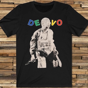 May include: Black t-shirt featuring a grayscale image of a person in a white outfit holding a musical instrument. The word "DEVO" is printed across the top in blue, green, and yellow letters. The shirt is made of a soft material.