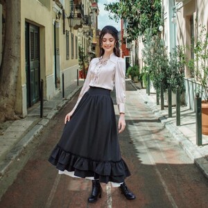 May include: A woman wearing a cream-colored blouse with a bow detail and a long black skirt with a ruffled hem. The skirt is paired with black boots. The outfit is styled in a vintage aesthetic, suitable for a variety of occasions.