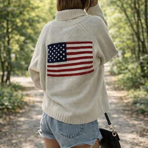 May include: A cream-coloured turtleneck sweater featuring a large American flag design on the back. The flag has a blue square with white stars and red and white stripes. The sweater is worn with denim shorts and a black handbag.