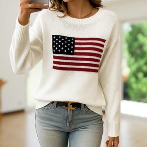 May include: White sweater featuring a knitted American flag design on the front. The flag has a black square with white stars and red and white stripes. The sweater has long sleeves and a crew neck.