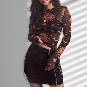 Pode incluir: Uma blusa preta de manga comprida em malha com um padrão celestial de sol, lua e estrelas em laranja. A blusa é combinada com uma minissaia preta franzida com zíper frontal. Um colar de ouro também é visível.