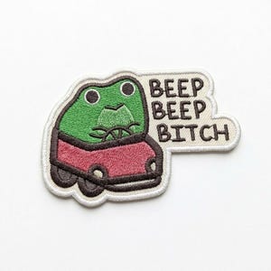 May include: Embroidered patch featuring a green frog driving a red car. The frog has large eyes and is behind the steering wheel. The patch includes the text "BEEP BEEP BITCH" in black letters.