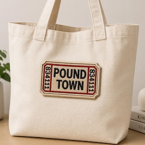 May include: A cream-colored tote bag with two handles. The bag features a rectangular patch with the words "POUND TOWN" in black and red, and the numbers "834111" on each side.