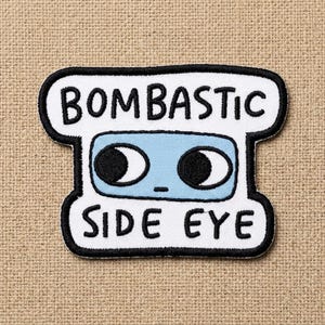 May include: Embroidered patch with the words "BOMBASTIC SIDE EYE" in black lettering. The patch features a light blue rectangle with two black eye shapes. The patch has a white background and a black border.