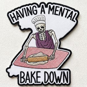 May include: Embroidered patch featuring a skeleton chef rolling dough with a rolling pin. The skeleton wears a chef's hat and apron. The patch has the text "HAVING A MENTAL BAKE DOWN" in black lettering on a white background with a black border.