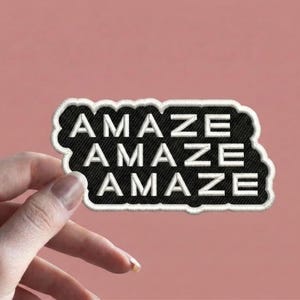 May include: A black embroidered patch with the word "AMAZE" repeated three times in white block letters. The patch has a white border and a black background. The patch is held against a pink background.