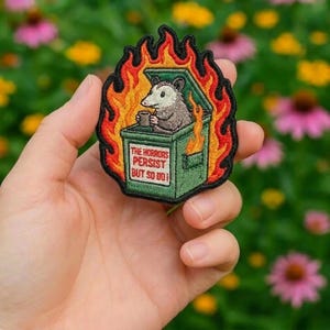 May include: Embroidered patch featuring an opossum in a green dumpster engulfed in flames. The opossum holds a mug. Text on the dumpster reads "THE HORRORS PERSIST BUT SO DO I!" The patch is outlined in black and surrounded by orange and red flames.