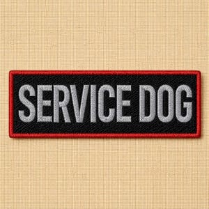 May include: Embroidered patch with the words "SERVICE DOG" in white capital letters on a black rectangular background. The patch has a red border and is set against a beige fabric.