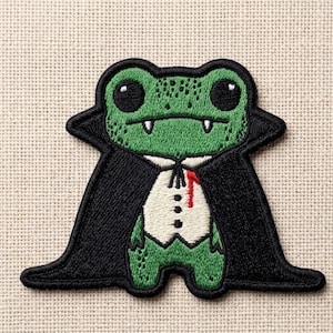 May include: Embroidered patch featuring a green frog dressed as a vampire. The frog has large black eyes, fangs, and is wearing a black cape and a white vest with a red accent. The patch is on a tan fabric background.