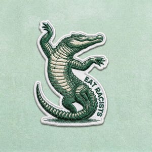May include: Embroidered patch featuring a green alligator in a dancing pose. The alligator has detailed scales and is outlined in white. The text "EAT RACISTS" is curved around the alligator's body in a matching green color.