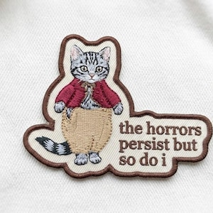 May include: Embroidered patch featuring a cat wearing a red jacket and tan pants. The cat has gray and black fur. Text on the patch reads "the horrors persist but so do i". The patch has a brown border.