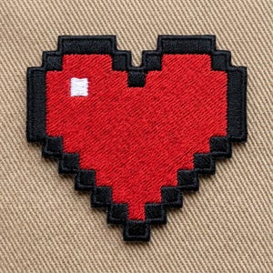 May include: Embroidered patch featuring a pixelated red heart with a black outline. A small white square is visible within the heart. The patch is likely for clothing or accessories.