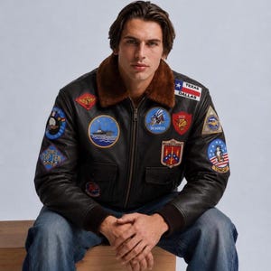 May include: A dark brown leather bomber jacket featuring a brown faux fur collar, ribbed cuffs, and numerous embroidered military-style patches on the chest and sleeves, including text reading Texas Dallas, Seabees, CVW-2 Top Ten, and Tomcat.