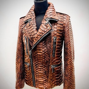 Op de afbeelding: Bruine leren jas met een slangenpatroon. De jas heeft een biker-stijl met een reverskraag, meerdere ritszakken en een diagonale ritssluiting. De jas wordt op een mannequin gepresenteerd.