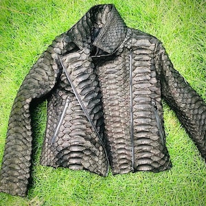 May include: A dark gray leather jacket with a textured, scale-like pattern. The jacket features a high collar, a diagonal zipper closure, and two zippered pockets. The jacket is lying on a bed of green grass.