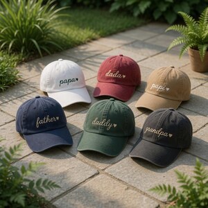 May include: Six baseball caps in various colors, including white, burgundy, tan, navy, green, and charcoal. Each cap features embroidered text: "papa", "dada", "papi", "father", "daddy", and "grandpa". The caps are arranged on a stone surface.