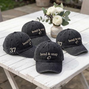 May include: Four charcoal grey baseball caps with a distressed finish. Each cap features the embroidered text "bend & snap" in a light beige font, accompanied by a small football icon. Two of the caps display the number "37" on the side panel.