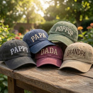 May include: A collection of six baseball caps in various colors, including black, navy, burgundy, olive green, and tan. Each cap features embroidered text: "PAPPY", "PAPA", "DADA", "POPOOP", and "GRANDPA". The caps are arranged on a wooden surface.