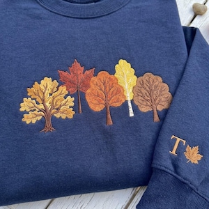 May include: A navy blue crewneck sweatshirt featuring an embroidered design of five distinct trees in autumn colors including gold, orange, and brown. The sleeve displays an embroidered letter T next to a small maple leaf in matching thread colors.