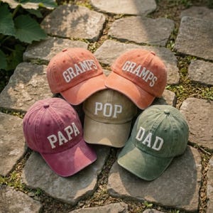 May include: Five baseball caps in various colors, including orange, burgundy, olive green, and tan. Each cap features white embroidered text: "GRAMPS", "PAPA", "POP", and "DAD". The caps are arranged on a stone surface.
