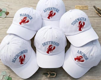 Peut inclure: Plusieurs casquettes de baseball blanches, ornées de broderies et d'un motif de homard rouge. Les mots brodés incluent "Adventure" et "Mariner". L'ensemble est présenté sur une surface en bois, évoquant un thème côtier.