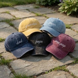 May include: Five baseball caps in various colors, including yellow, blue, and burgundy. Each cap has a different embroidered word: "grandpa", "papa", "dad", "pops", and "pap pap". The caps are arranged on a stone surface.