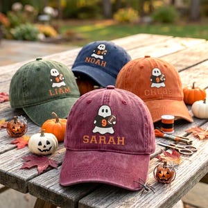 May include: Four baseball caps in olive green, navy blue, orange, and maroon. Each cap features an embroidered ghost wearing a football jersey with the number 9 and a football, with the names Liam, Noah, Chloe, and Sarah stitched below.