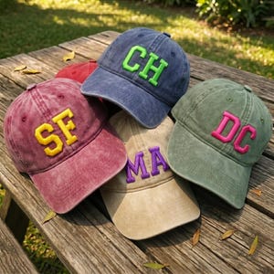 May include: A collection of baseball caps in various colors, including burgundy, navy, khaki, and olive green. Each cap features embroidered letters in contrasting colors, such as yellow, green, pink, and purple. The caps are arranged on a wooden surface.