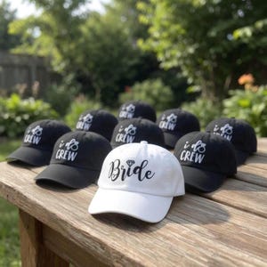 May include: A collection of black baseball caps with the text "I do Crew" in white, and one white cap with "Bride" in black script and a diamond graphic. The caps are arranged on a wooden surface.