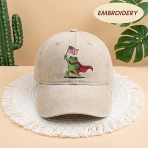 May include: Tan baseball cap featuring an embroidered frog superhero holding an American flag. The frog wears a red cape. The word "EMBROIDERY" is printed on the top right of the cap.