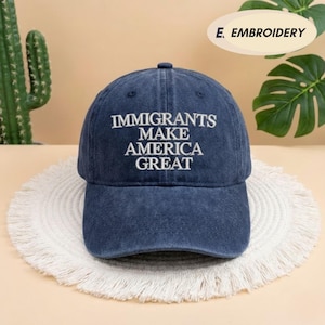 May include: A navy blue baseball cap with the embroidered text "IMMIGRANTS MAKE AMERICA GREAT" in white. The cap is made of a soft fabric and has a curved brim. The cap is sitting on a white, fringed placemat.