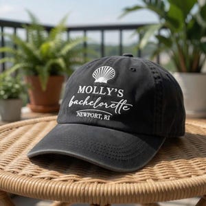 May include: A black baseball cap with a curved brim. The cap features the text "MOLLY'S bachelorette NEWPORT, RI" in white script, along with a seashell graphic. The hat is sitting on a wicker table.